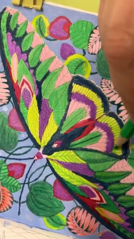 Butterfly with leaf-patterned wings in lime yellow, purple and pink among green and coral leaves in Millie Marotta's Curious Creatures, being colored with a green colored pencil